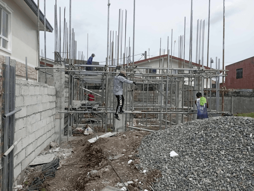 Construction site with scaffolding, workers, and building materials in front of residential houses.